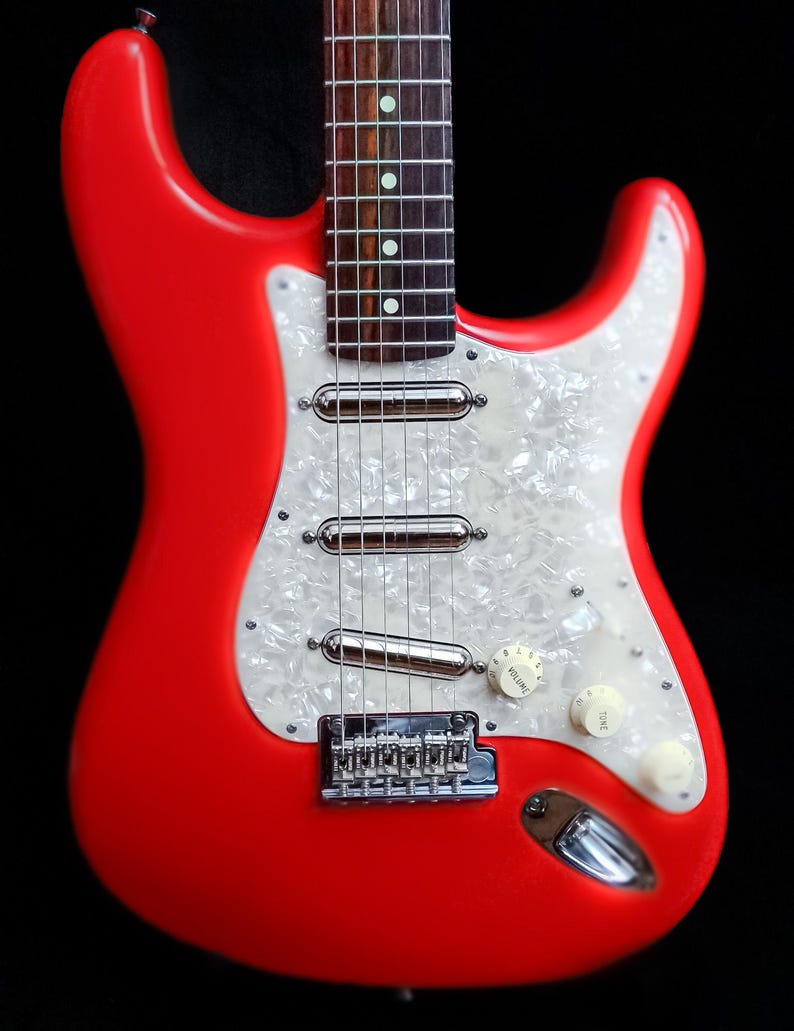 Pu&ograve; includere: Una vivace chitarra elettrica rossa con battipenna perlato. La chitarra presenta hardware cromato, inclusi pickup, ponte e meccaniche. Sono visibili manopole di controllo bianche. Il corpo della chitarra &egrave; rosso pieno e lucido.