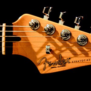 Pu&ograve; includere: Primo piano della paletta di una chitarra Fender Stratocaster. La paletta &egrave; in legno chiaro con il logo Fender e il testo Stratocaster. Le meccaniche cromate e le corde sono visibili su sfondo nero.