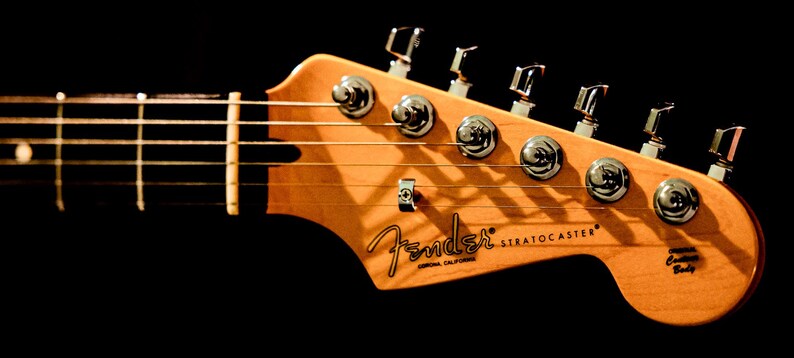 Pu&ograve; includere: Primo piano della paletta di una chitarra Fender Stratocaster. La paletta &egrave; in legno chiaro con il logo Fender e il testo Stratocaster. Le meccaniche cromate e le corde sono visibili su sfondo nero.
