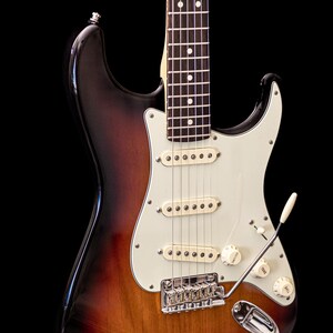 Pu&ograve; includere: Chitarra elettrica sunburst con battipenna bianco, tre pickup e hardware cromato. La chitarra ha un manico in acero e un braccio tremolo. Il corpo &egrave; una sfumatura di marrone, arancione e nero.