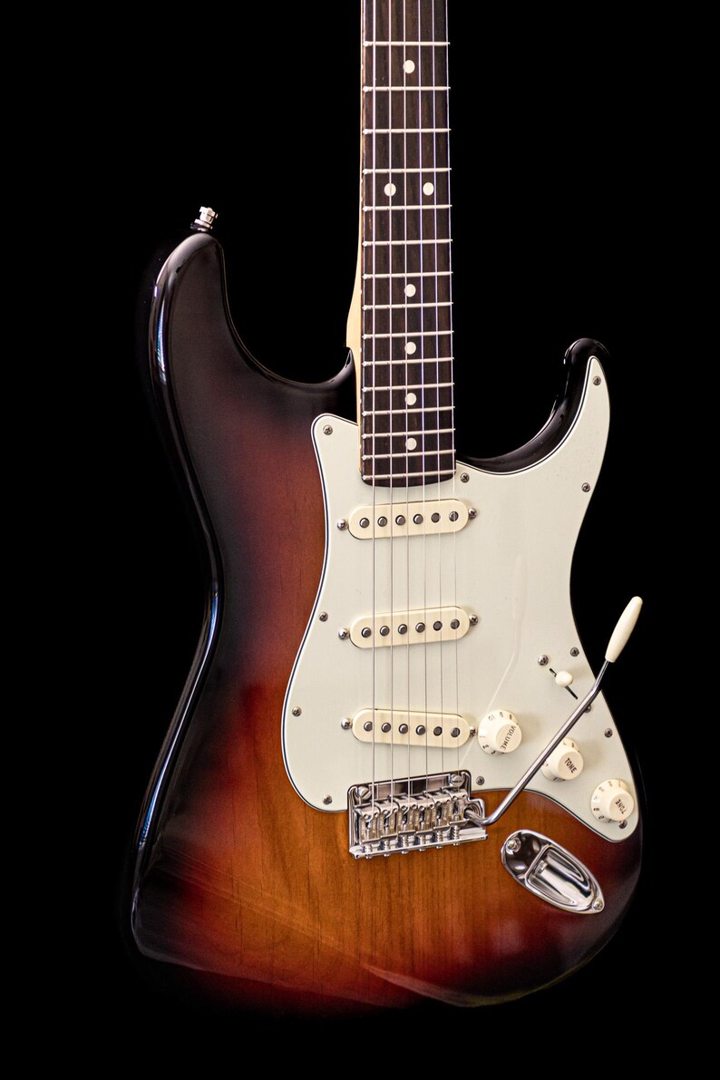 Pu&ograve; includere: Chitarra elettrica sunburst con battipenna bianco, tre pickup e hardware cromato. La chitarra ha un manico in acero e un braccio tremolo. Il corpo &egrave; una sfumatura di marrone, arancione e nero.