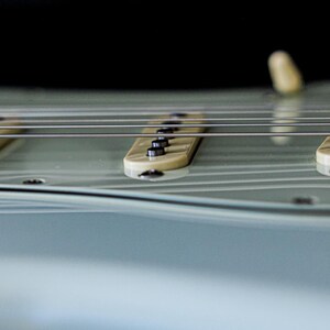 Pu&ograve; includere: Primo piano di una chitarra elettrica con corpo azzurro. L'immagine mette in evidenza le corde, i pickup e le manopole di controllo. Il design della chitarra &egrave; elegante, con un focus sull'hardware dello strumento e la curva del corpo.