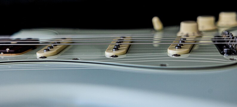 Pu&ograve; includere: Primo piano di una chitarra elettrica con corpo azzurro. L'immagine mette in evidenza le corde, i pickup e le manopole di controllo. Il design della chitarra &egrave; elegante, con un focus sull'hardware dello strumento e la curva del corpo.