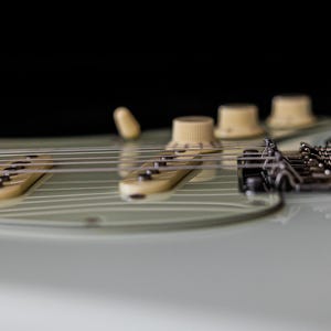 Pu&ograve; includere: Primo piano di una chitarra elettrica, che mostra le corde, i pickup e le manopole di controllo. La chitarra ha un corpo verde chiaro con pickup e manopole color crema. Le corde sono argentate, su sfondo nero.