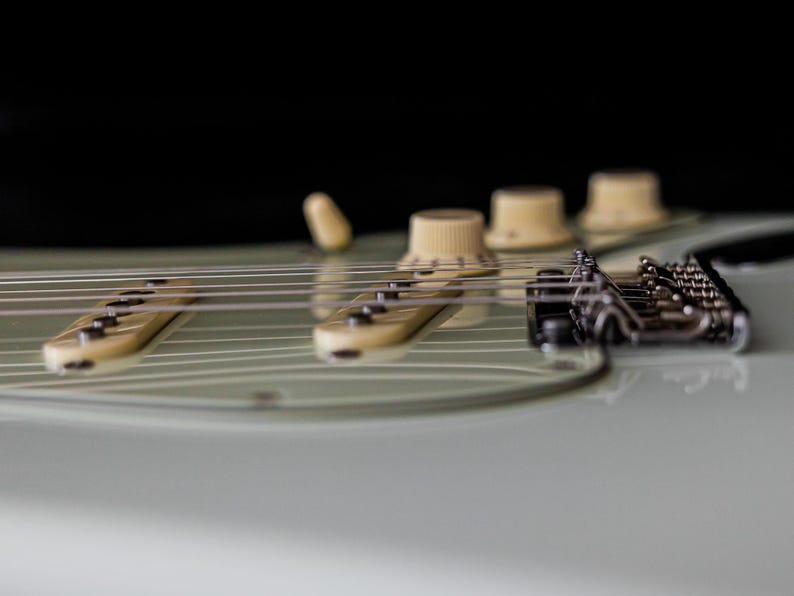 Pu&ograve; includere: Primo piano di una chitarra elettrica, che mostra le corde, i pickup e le manopole di controllo. La chitarra ha un corpo verde chiaro con pickup e manopole color crema. Le corde sono argentate, su sfondo nero.