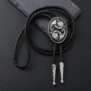 May include: A black leather bolo tie with an oval silver-toned centerpiece. The centerpiece features a floral design with intricate detailing. The tie has silver-toned tips and a braided leather cord. The bolo tie is a Western-style accessory.