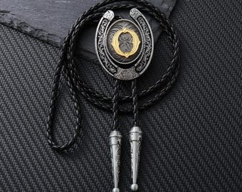 Könnte beinhalten: Eine silber-goldene Bolo-Krawatte mit einer schwarzen geflochtenen Kordel. Die Bolo-Krawatte hat einen ovalen Anhänger mit einem verzierten Design und einem goldenen Buchstaben "O" in der Mitte. Das Kordelende hat silberfarbene Zierspitzen.