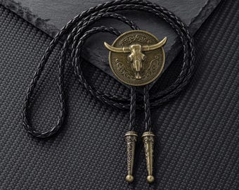 Puede incluir: Una corbata bolo con un colgante circular de color latón antiguo con un diseño de calavera de toro. El colgante está unido a un cordón de cuero trenzado negro con puntas metálicas decorativas.
