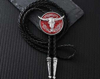 Puede incluir: Una corbata bolo con un colgante de calavera de toro plateado sobre un fondo rojo, detallado con detalles plateados. El cordón de cuero trenzado negro tiene puntas plateadas. Un accesorio de estilo Western.