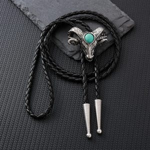 May include: A silver-colored ram head bolo tie with a turquoise stone set in the center. The black braided leather cord has silver-colored tips and a sliding clasp. The bolo tie is on a black textured surface.