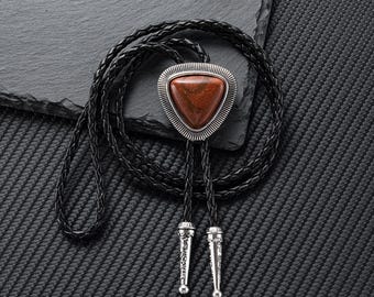 May include: A bolo tie featuring a triangular, reddish-brown stone set in a silver-colored frame. The tie has a braided black leather cord and decorative silver-colored tips. The accessory is displayed on a textured black surface.