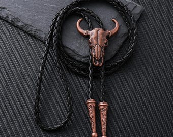 Puede incluir: Una corbata bolo con un colgante de calavera de toro color cobre. El colgante está unido a un cordón de cuero negro trenzado. El cordón tiene puntas decorativas de color cobre. La corbata se muestra sobre una superficie oscura texturizada.