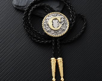 May include: A bolo tie with a circular silver and gold pendant featuring an ornate design and the letter "C". The black braided leather cord has gold-colored tips. The tie rests on a black textured surface.