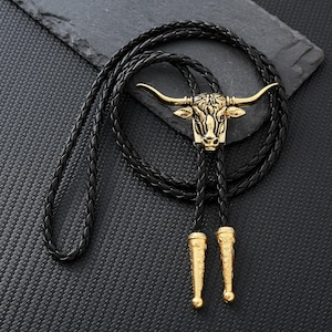Peut inclure: Une cravate bolo en cuir noir avec un pendentif tête de taureau doré. Le pendentif présente des cornes et des traits du visage détaillés. La cravate a un design tressé et des embouts dorés.