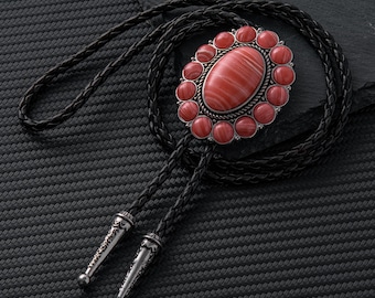 May include: A bolo tie with a large oval red stone center, surrounded by smaller red stones and a silver-colored metal frame. The black leather cord has decorative silver tips. The tie is set against a dark background.