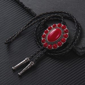 May include: A bolo tie featuring a large oval red stone centerpiece surrounded by smaller red stones and a silver-colored metal frame. The black braided leather cord has decorative silver-colored metal tips.