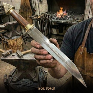 Könnte beinhalten: Ein handgefertigter Dolch mit Holzgriff und Messingakzenten. Die Klinge weist ein geätztes Design auf. Das Bild zeigt den Dolch in einer Schmiede, mit dem Text "RÖK FÖRGE" unten.