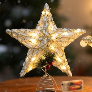 Könnte beinhalten: Zwei sternförmige Weihnachtsbaumspitzen. Jeder Stern ist weiß mit goldenen Akzenten und wird von kleinen Lichtern beleuchtet. Die Basis ist eine silberne Spirale mit einem kleinen Zweig Grün und roten Beeren. Ein Batteriepack ist sichtbar.