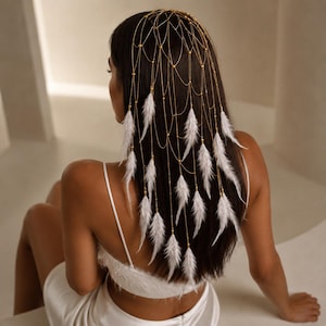 May include: A gold-toned metal headpiece featuring a delicate web-like chain structure draped over the hair. The accessory is adorned with small gold beads and multiple white feathers hanging from the chains, creating a layered, cascading effect.