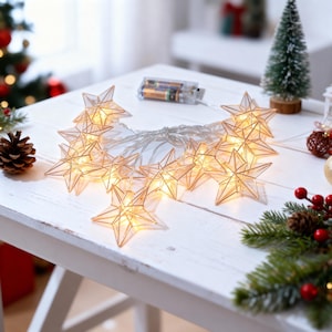 Pode incluir: Uma sequência de luzes em forma de estrela com iluminação branca quente. As estrelas de plástico transparente são conectadas por um fio fino e prateado e alimentadas por um compartimento de bateria. Decorações de Natal em uma superfície branca.