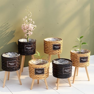 May include: A collection of woven planters with wooden legs. The planters are in various shades of brown and tan, some holding plants with green leaves and pink flowers. Each planter has a label that reads "Natural Home Flower".