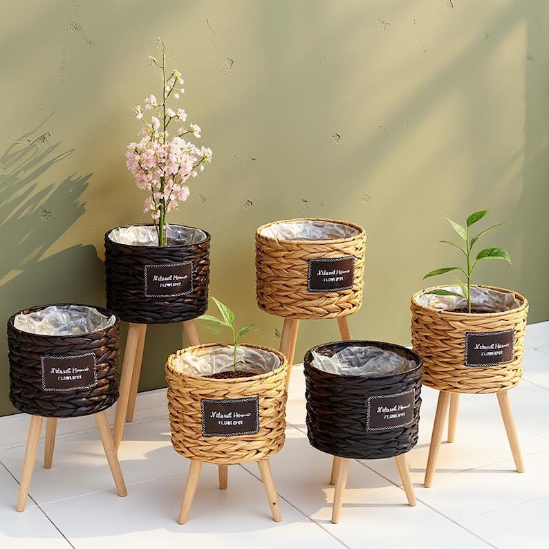 May include: A collection of woven planters with wooden legs. The planters are in various shades of brown and tan, some holding plants with green leaves and pink flowers. Each planter has a label that reads "Natural Home Flower".
