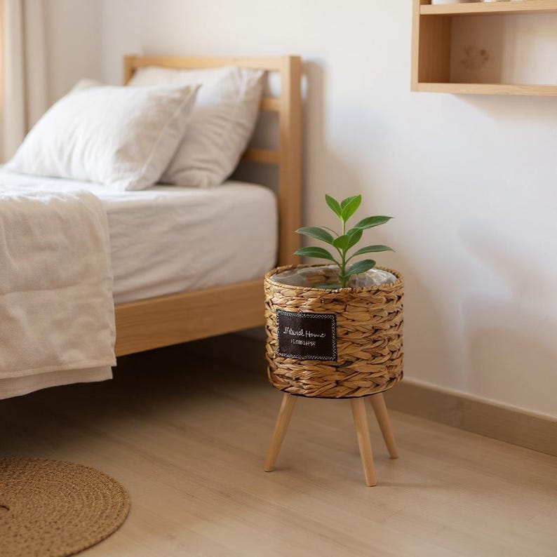 May include: A woven wicker planter with a small green plant, set on three wooden legs. The planter has a dark rectangular label with white text. The planter is near a bed with white linens.