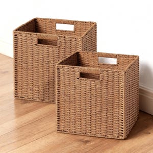 May include: Two woven brown storage baskets with rectangular openings and built-in handles. The baskets are different sizes and sit on a wooden floor. The baskets are made of a woven material.