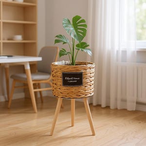 Könnte beinhalten: Ein geflochtener Weidenkorb-Pflanztopf mit einer Monstera-Pflanze, auf einem dreibeinigen Holzständer. Der Topf hat ein schwarzes Etikett mit der Aufschrift "Natural Home I LOVE SPOT". Die Pflanze hat große, grüne, herzförmige Blätter.