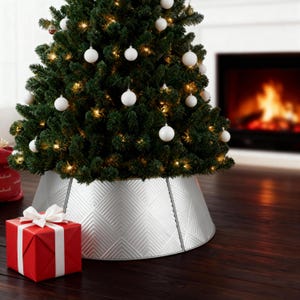 Op de afbeelding: Een versierde kerstboom met witte ballen en lichtjes, omringd door een zilveren boomkraag met een geometrisch patroon. Een verpakte rode geschenkdoos met een witte strik staat in de buurt. De scène speelt zich af in een kamer met een open haard.