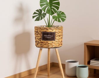May include: A woven basket planter with a Monstera plant, set on a wooden tripod stand. The planter has a brown label that reads "Natural Home FLOWER SPOT". Two additional planters are visible in the background.