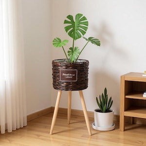 May include: A brown woven planter with a plant, set on a three-legged wooden stand. The planter has a rectangular label with the text "Natural Home I PRESENT". A smaller white potted plant sits nearby.