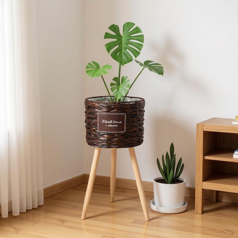 May include: A brown woven planter with a plant, set on a three-legged wooden stand. The planter has a rectangular label with the text "Natural Home I PRESENT". A smaller white potted plant sits nearby.