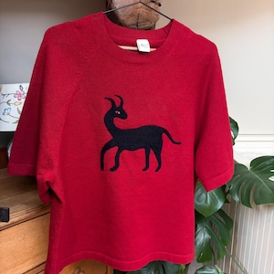 May include: A red short-sleeved sweater with a black animal design. The sweater is hanging on a black hanger. The sweater is made of a soft material. The design is a black silhouette of an animal with horns.