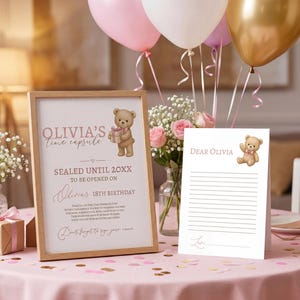 以下が含まれることがあります： ピンク色のテーブルクロスの上に、額入りのサインとカードがあります。「Olivia's time capsule」と「Sealed until 20XX」と書かれたサイン。テディベアのイラストがサインとカードの両方にあります。背景には風船と花が飾られています。