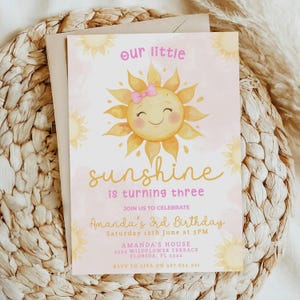 May include: A birthday invitation featuring a smiling sun illustration with a pink bow. The text reads "our little sunshine is turning three" and includes party details. The invitation is set against a light pink background.