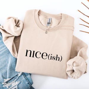May include: A beige sweatshirt with the text "nice(ish)" printed in black. The sweatshirt is folded with the sleeves tucked in. A silver necklace with a small pendant is draped over the top. Blue denim jeans are visible in the background.