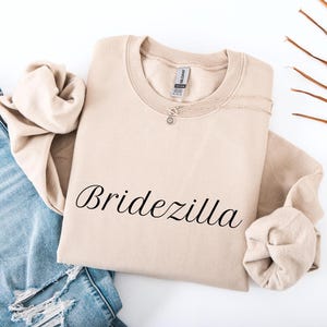 Puede incluir: Una sudadera beige con la palabra "Bridezilla" en escritura negra. Un collar plateado con un pequeño colgante descansa sobre la camisa. La sudadera está doblada junto a unos vaqueros azules.