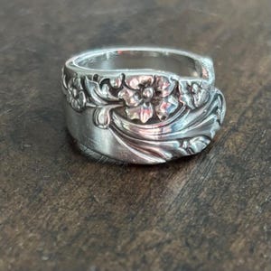 May include: A silver-toned metal ring crafted from a repurposed spoon, featuring an embossed floral pattern with detailed petals and leaves that wrap around the band. The surface has a polished finish with darkened recessed areas for contrast.