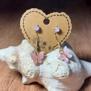 May include: A pair of pink butterfly earrings with gold-colored hooks and a small, light purple gem at the top. The earrings are displayed on a brown, heart-shaped tag with a floral design, resting on a seashell.