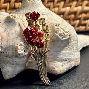May include: A gold-toned metal brooch featuring a bouquet of red flowers. The brooch has a vertical design with detailed leaves and stems. The flowers are a vibrant red color, and the brooch is set against a neutral background.