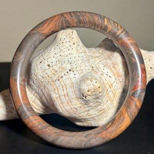 May include: A marbled bangle bracelet with shades of brown, orange, and gray. The bracelet has a smooth, polished finish and a circular shape. The bangle is resting on a white seashell.