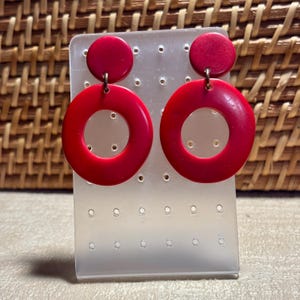 May include: A pair of bright red, retro-style earrings. Each earring features a small, circular stud connected to a large, hollow circle. The earrings are displayed on a clear, plastic earring holder against a woven background.