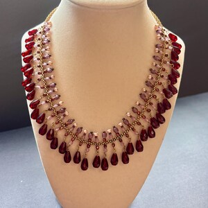May include: A beaded necklace featuring a gold-toned chain base adorned with dangling deep red teardrop-shaped beads and smaller light purple and pink spherical beads. The fringe-style design creates a layered effect with alternating bead sizes.
