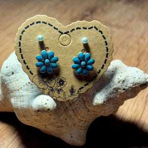 May include: A pair of turquoise flower stud earrings displayed on a heart-shaped card. The earrings have a silver-colored metal outline and a turquoise enamel flower design. The card is brown with a dotted border and floral illustrations.