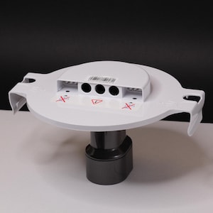 May include: A white plastic component with a circular top and a dark gray base. The top features three circular holes and a rectangular section with a barcode. Red "X" symbols and a warning symbol are also visible.