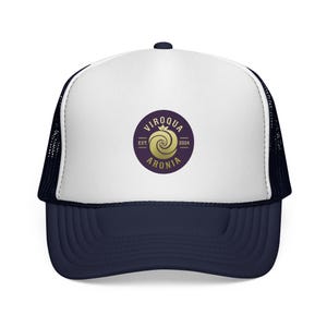 May include: A trucker hat with a white front panel and a navy blue brim and bill. The hat features a circular logo with the text "VIROQUA ARONIA" and a gold-colored spiral design. The hat has a mesh back.
