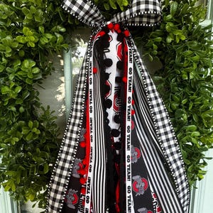 May include: A decorative wreath attachment featuring a black and white buffalo check bow with long cascading ribbons. The ribbons include red, white, and black patterns, red pom-pom trim, and text that reads GO TEAM! alongside Ohio State logos.