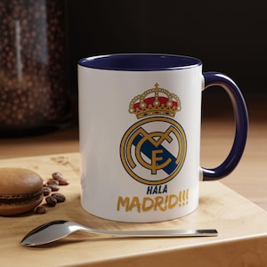 Puede incluir: Taza de cerámica blanca con interior y asa azules. Muestra el logo del Real Madrid en oro y azul, coronado por una corona, y las palabras "Hala Madrid!!!" en oro. Una cuchara y un macaron están sobre una superficie de madera.