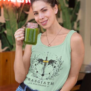 May include: A pale green vest top with a black dragon and moon graphic, and the text "BASGIATH WAR COLLEGE". The top is worn with blue jeans and a black belt. The person is holding a glass jar filled with green liquid.